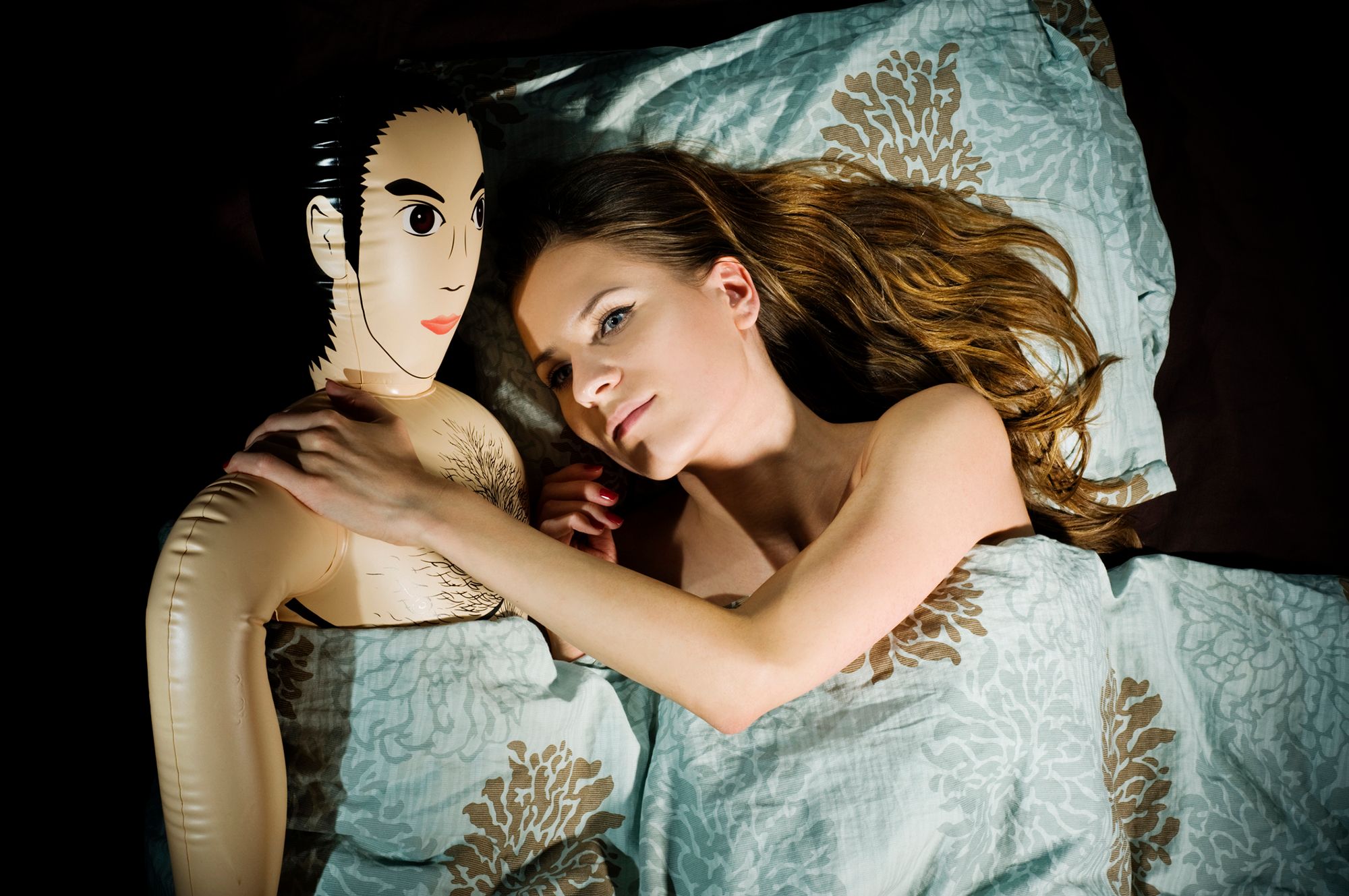 Frau liegt mit einer Männer-Gummipuppe im Bett