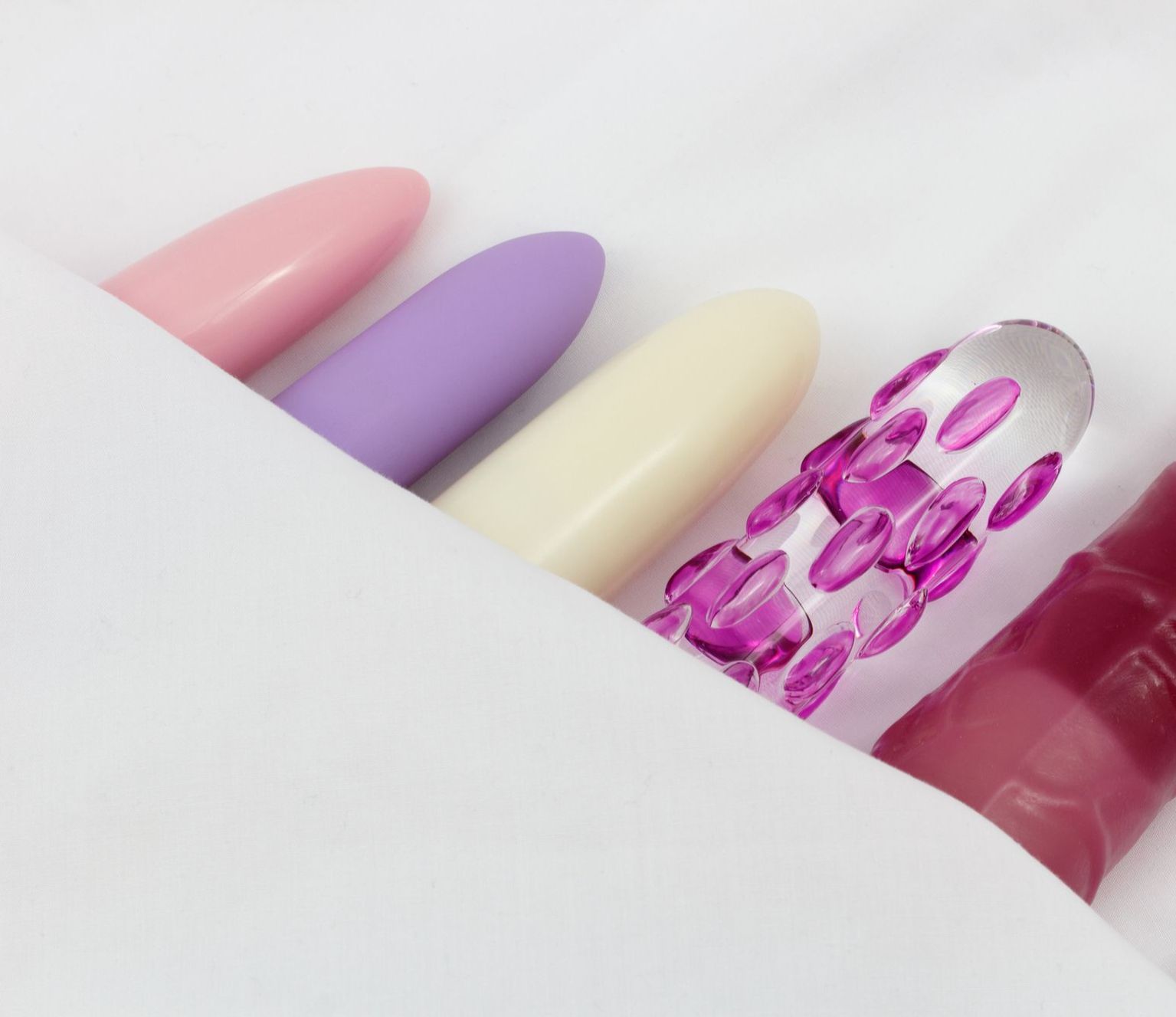 Rosaner, lilaner und gelber Vibrator sowie ein Glasvibrator und roter Naturvibrator unter einem Tuch auf weißem Untergrund