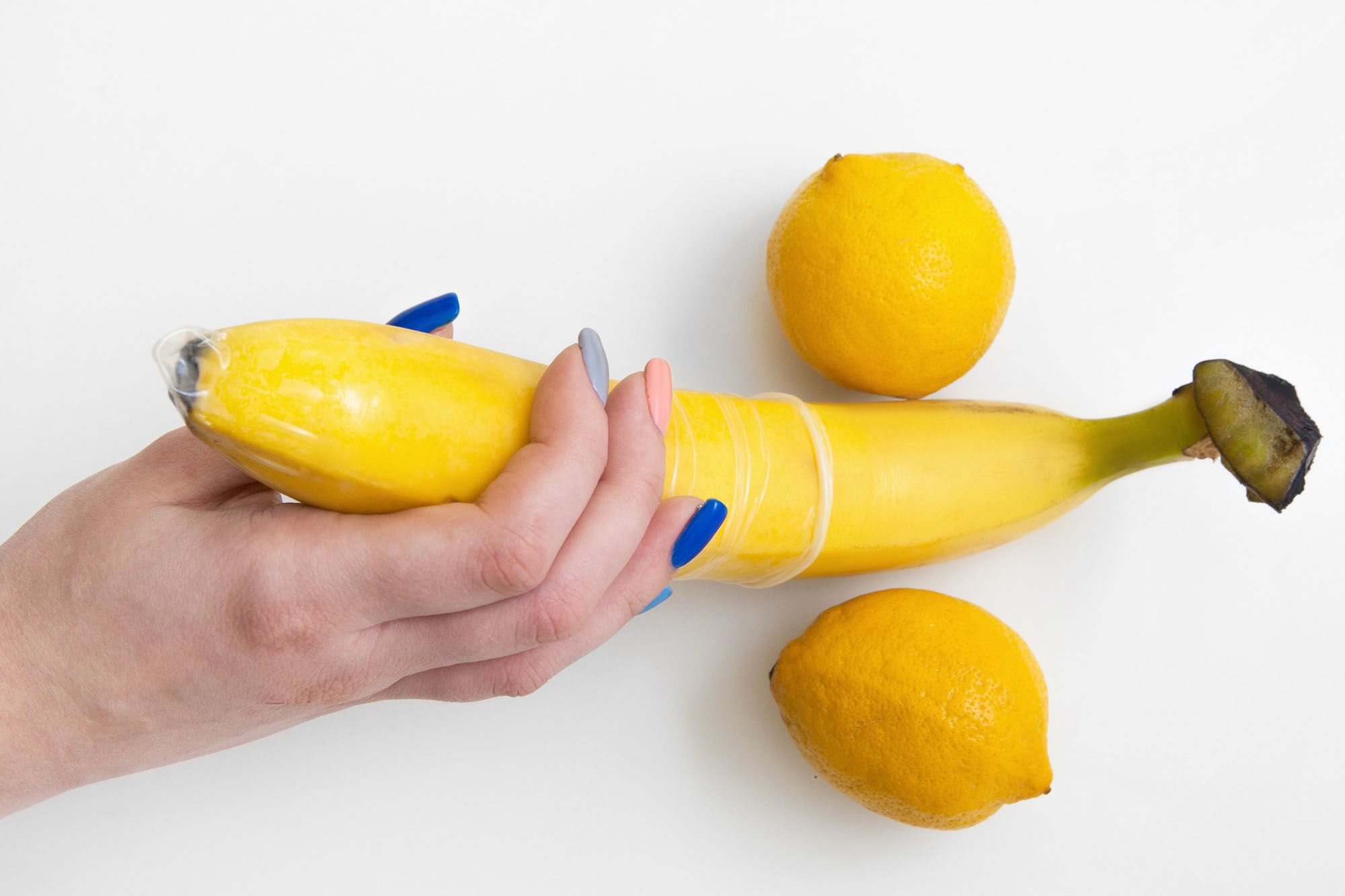 Hand hält eine Banane, über die ein Kondom gezogen ist, zwei Zitronen symbolisieren die Hoden