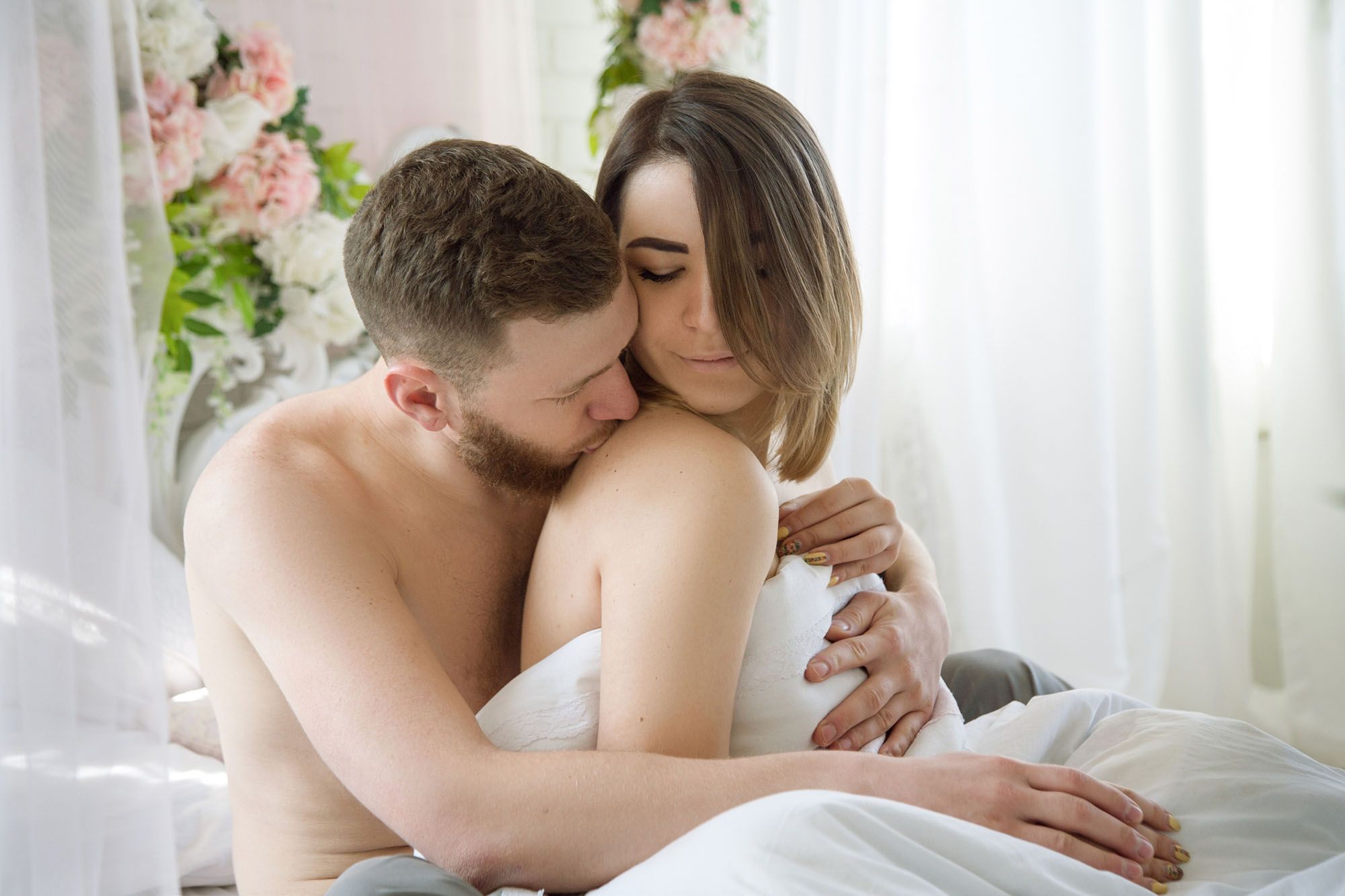 Mann sitzt hinter seiner Partnerin im Bett uns küsst ihre Schulter