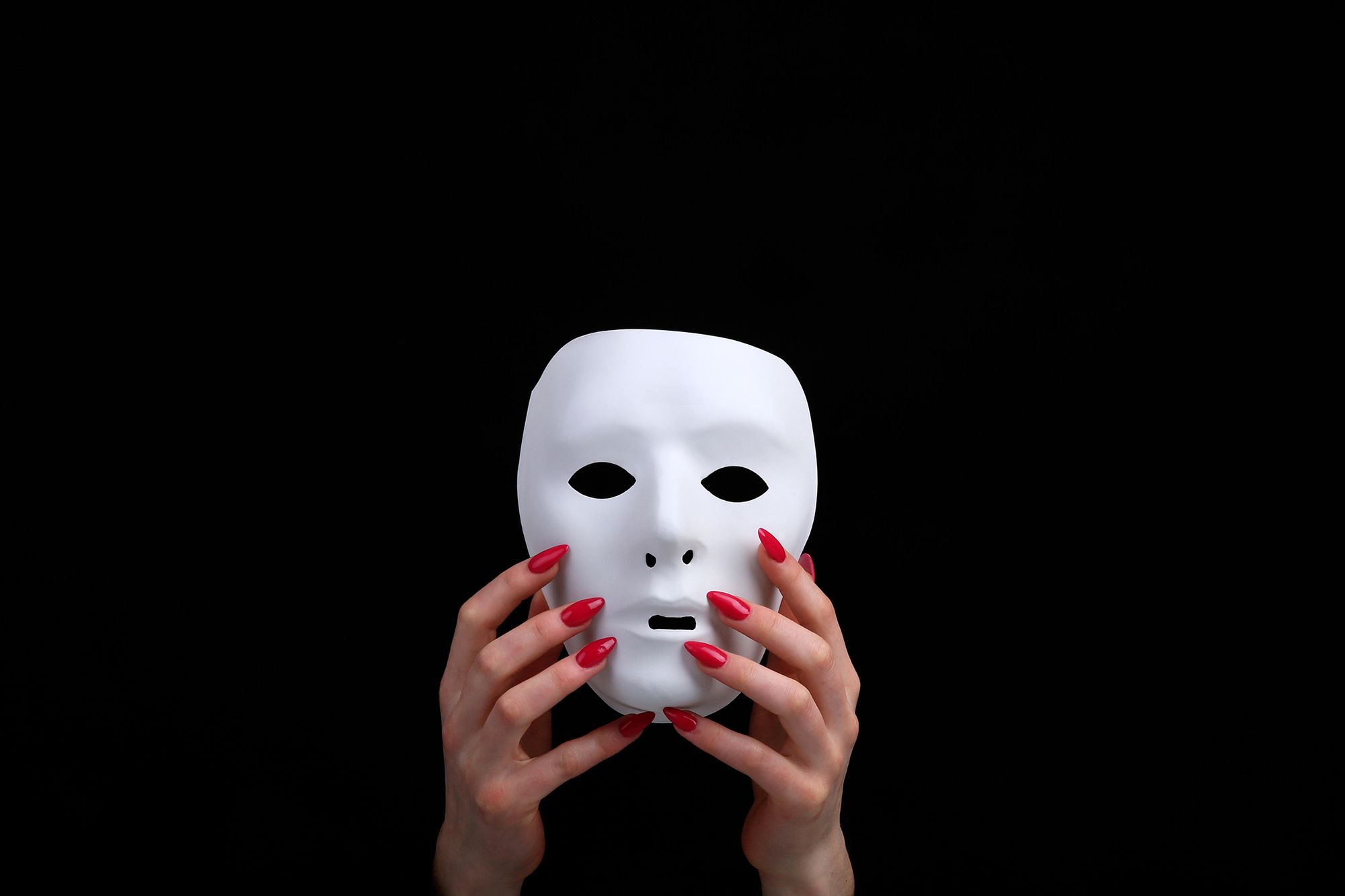 Hände mit roten langen Nägeln halten eine weiße Maske vor schwarzem Hintergrund