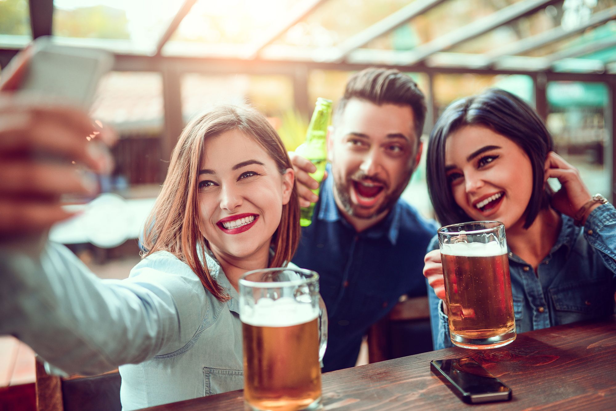 Brünette und schwarzhaarige Frau und brünetter Mann, die in einer Bar Bier trinken und ein Selfie schießen