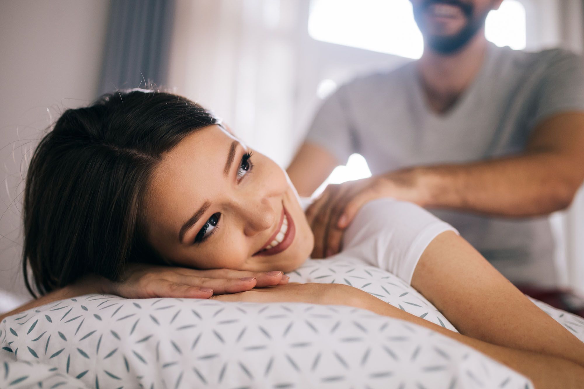 Frau in weißem T-Shirt, die auf dem Bauch auf einem Bett liegt und von einem Mann eine Rückenmassage erhält