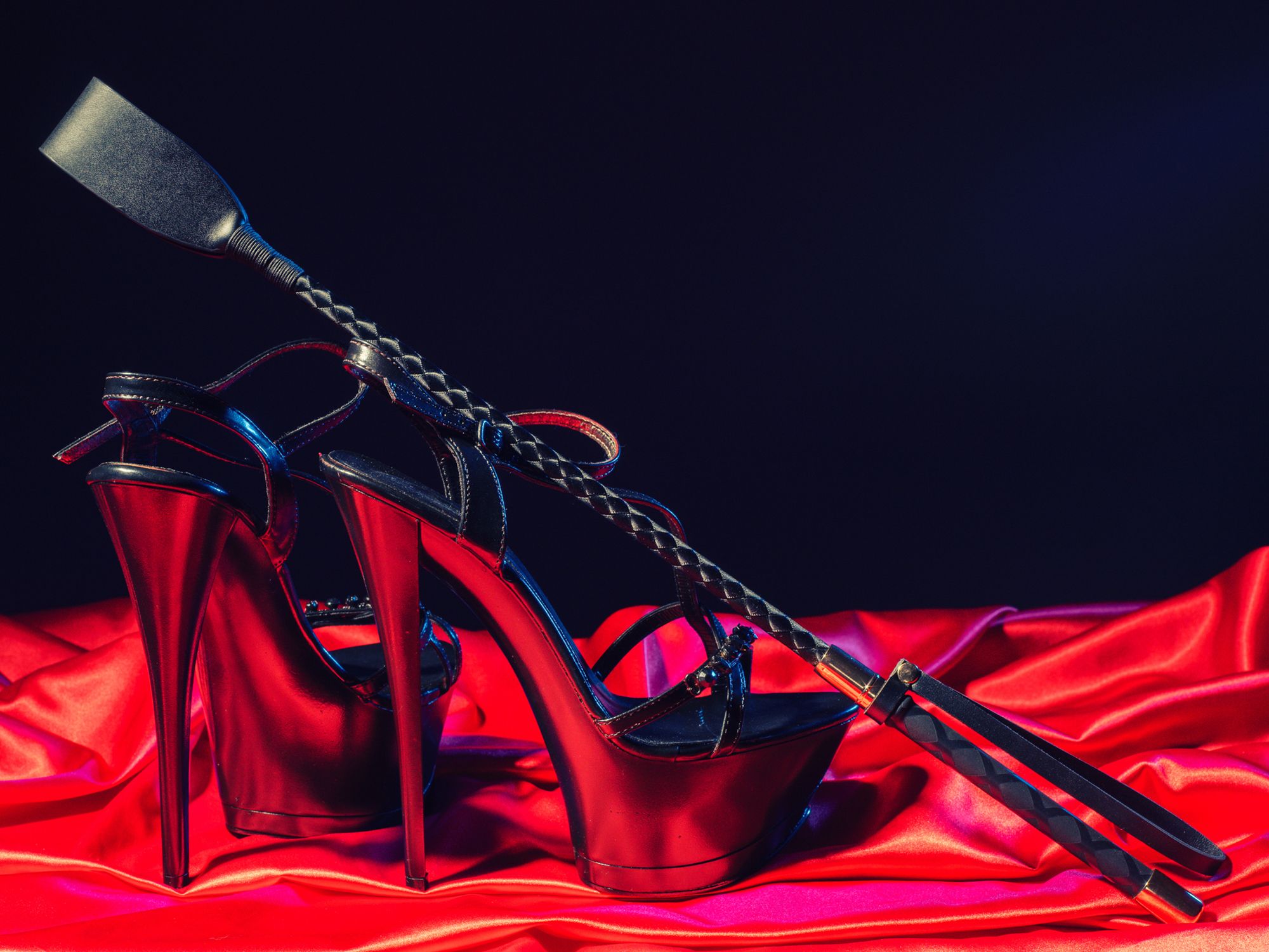 Schwarze High Heels und Reitgerte auf rotem Satinstoff
