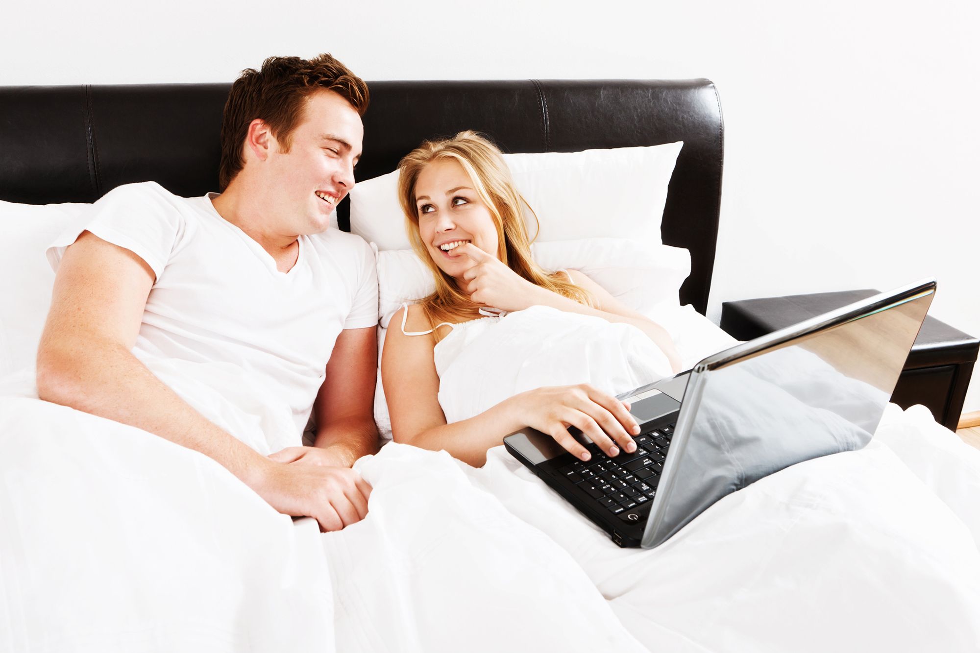 Blonde Frau und brünetter Mann, die nebeneinander mit einem Laptop auf dem Schoß im Bett liegen während die Frau verschmitzt zum Mann schaut