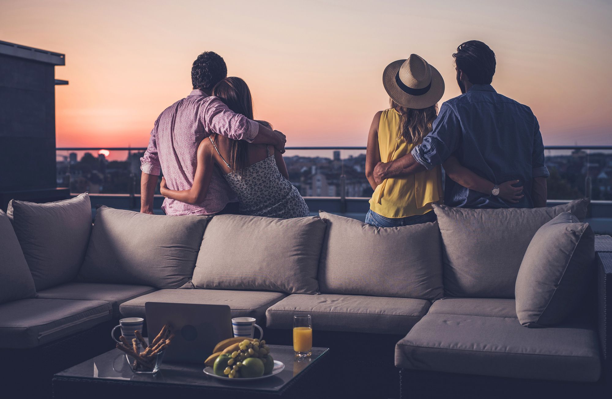 Partnertausch mit Freunden - Abbildung zweier sich umarmender Hetero-Paaren, welche nebeneinander auf einer Dachterrasse sitzen und im Sonnenuntergang über die Stadt blicken.