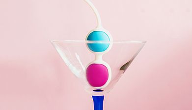 Ein weiß, pink und türkisfarbener Analplug der in einem Cocktailglas liegt vor rosanem Hintergrund