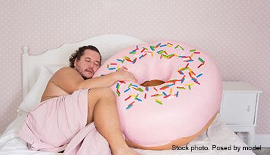 Ein Mann der im Bett mit einem übergroßen rosanem Donut kuschelt.