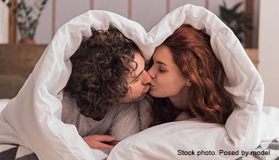 Ein Paar das in einem Bett liegt mit der Decke über den Köpfen und sich küsst.
