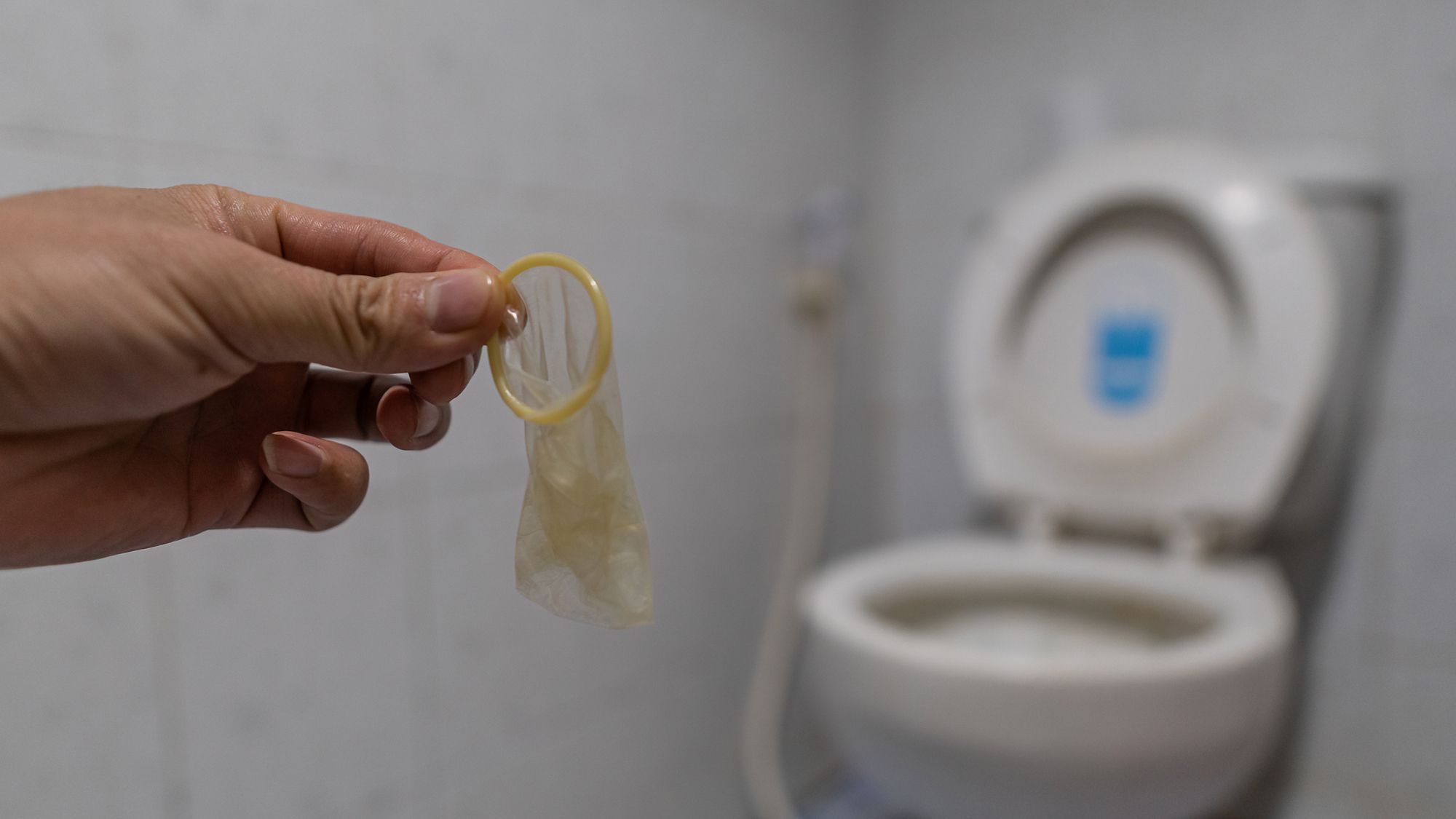 Eine Hand die ein benutztes Kondom hält, im Hintergrund ist eine Toilette