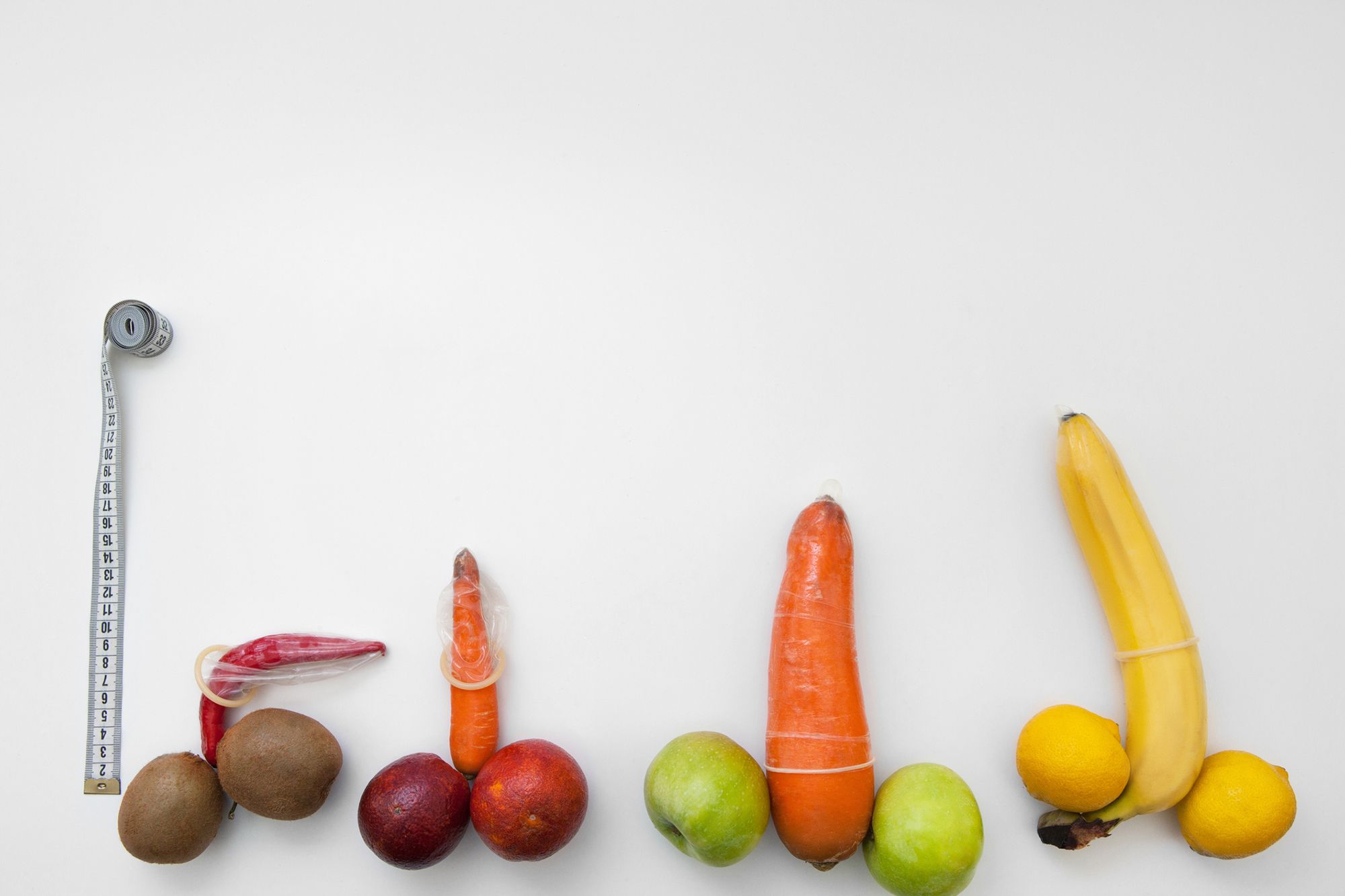 Obst und Gemüse in Penisform mit übergezogenen Kondomen neben Maßband