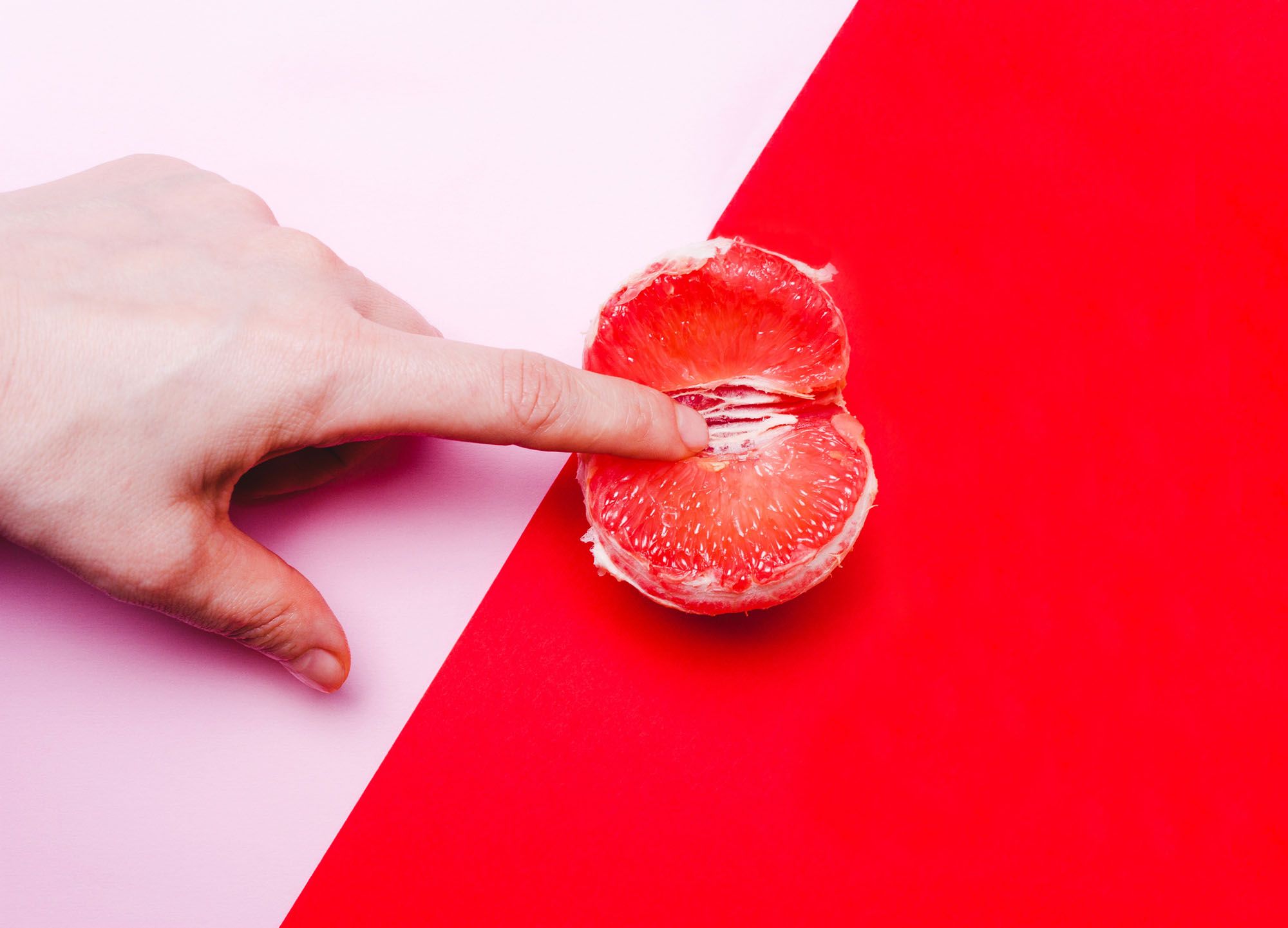 Finger, der in eine rote Grapefruit gesteckt wird auf einem rosa-rot geteilten Hintergrund