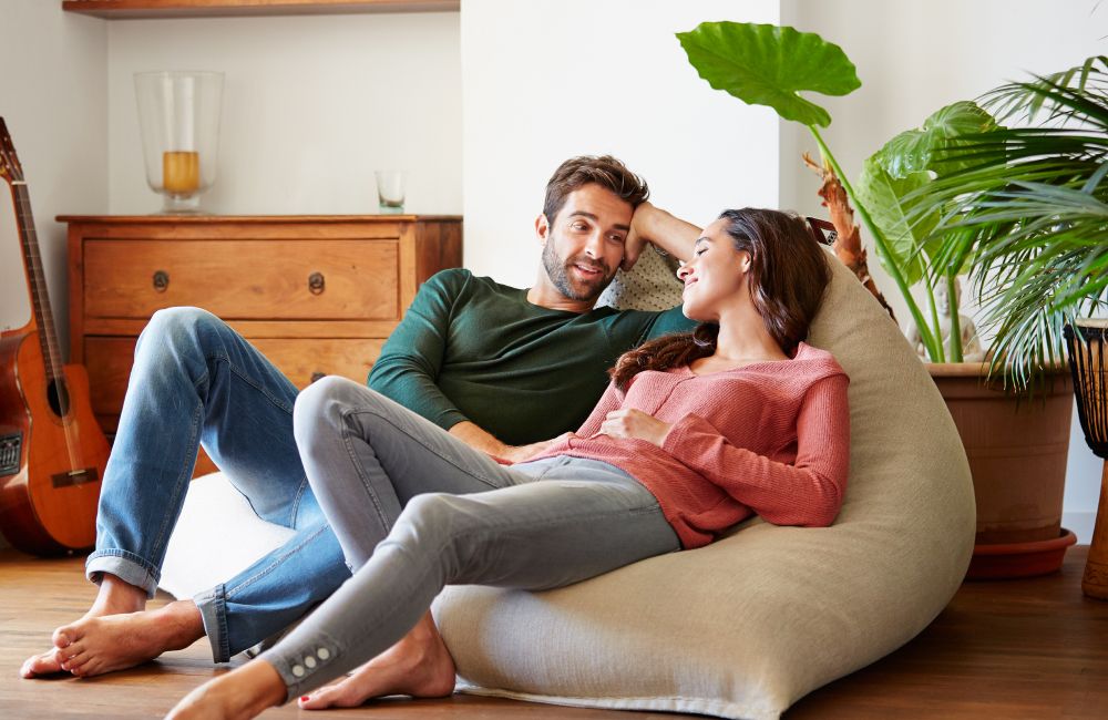 Frau in rotem Oberteil und grauer Jeans und Mann in grünem Sweatshirt und blauer Jeans, die auf einem Sitzsack in einem Wohnzimmer liegen