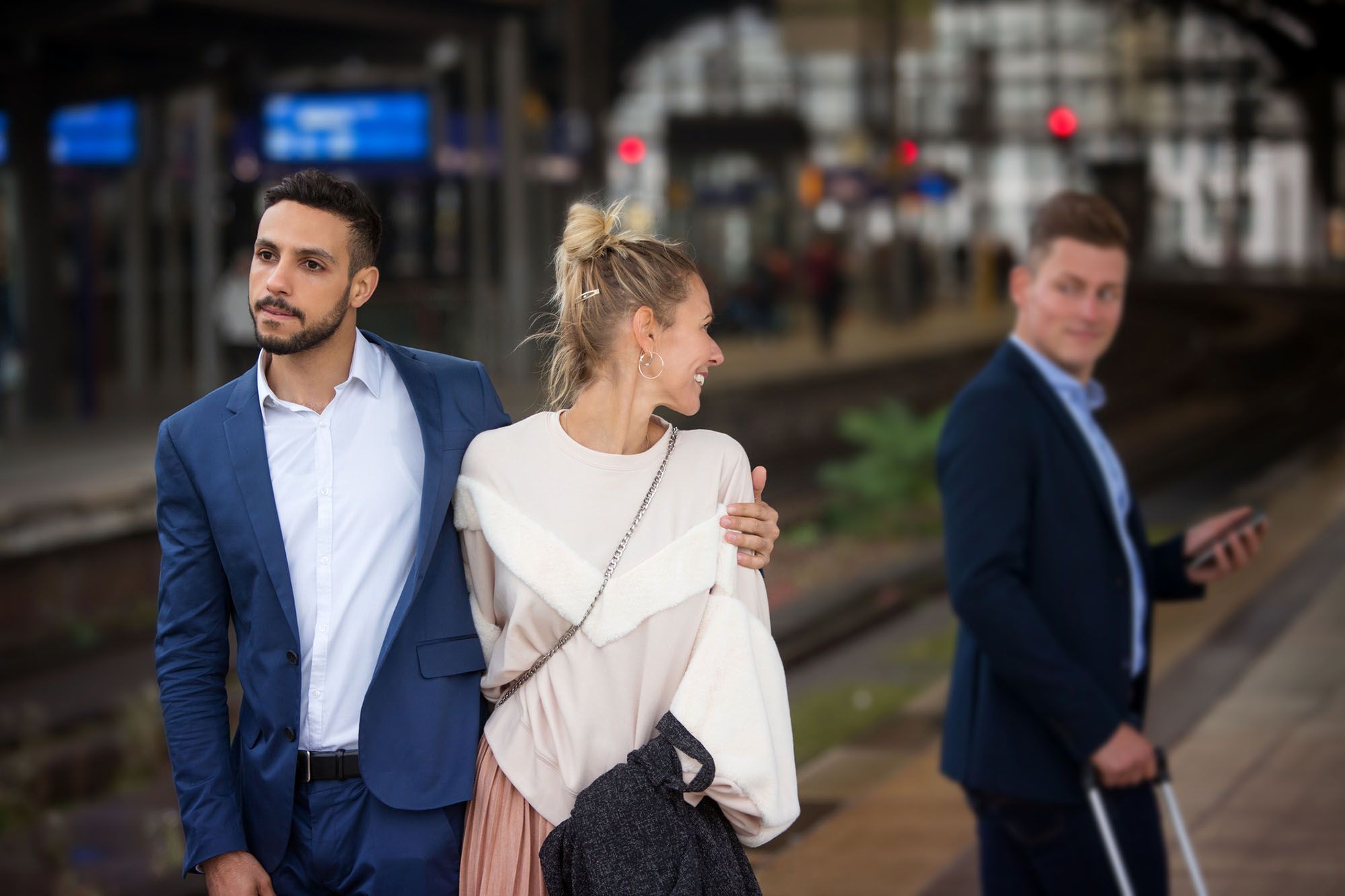 Ein Pärchen am Bahnhof. Der Mann hat seinen Arm um die Frau gelegt. Sie dreht sich um und lächelt einen anderen Mann an, der interessiert zurückblickt.