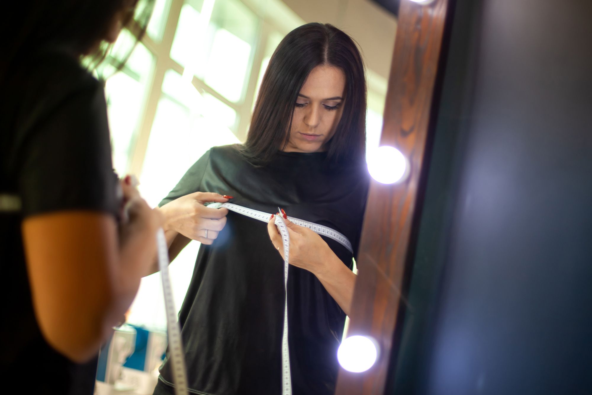 Eine Frau mit braunen Haaren steht vor einem Spiegel und legt ein Maßband um ihre Oberweite, um ihren Brustumfang zu messen