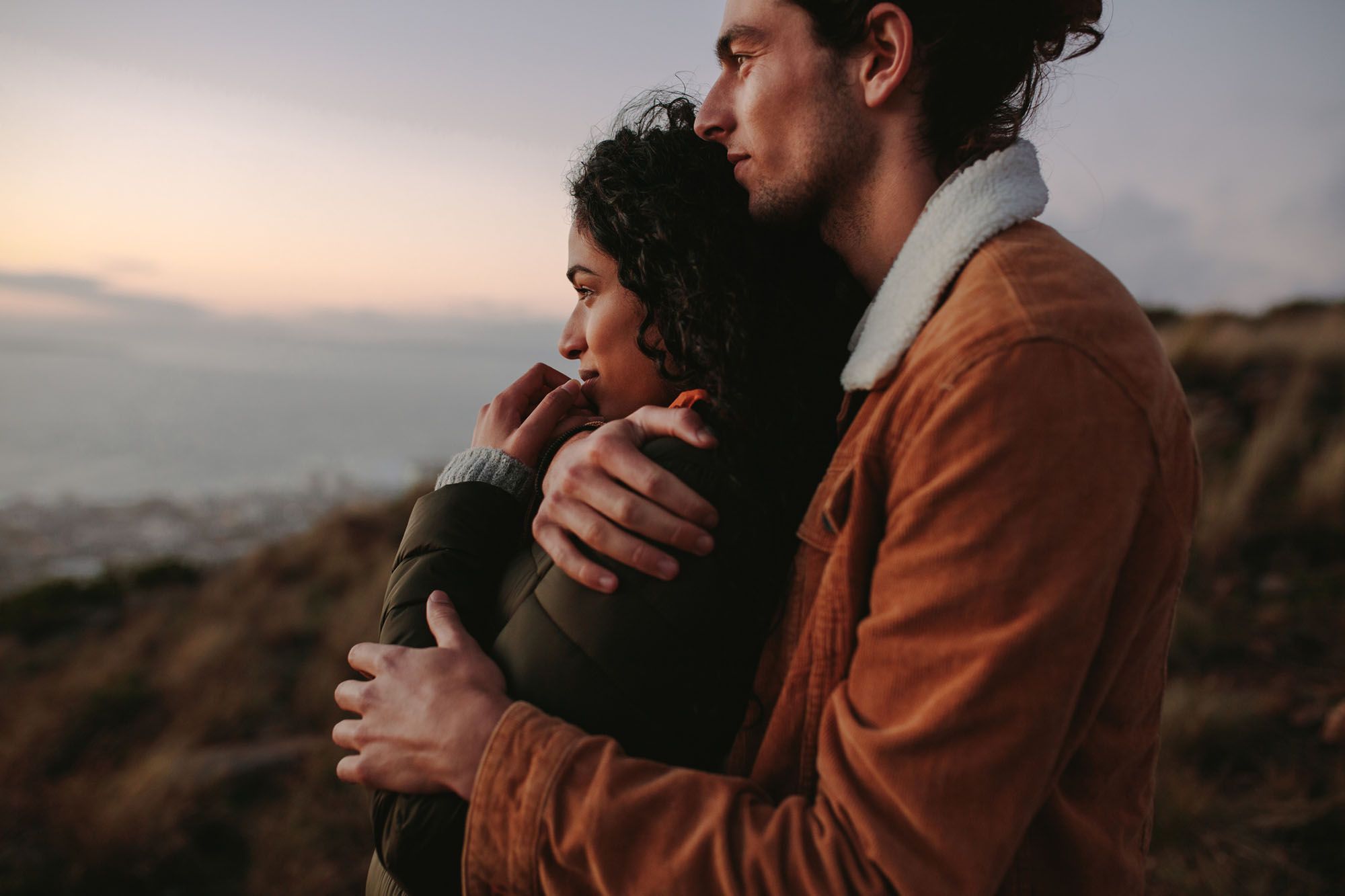 Mann in brauner Jacke umarmt Frau von hinten während beide den Sonnenuntergang betrachten