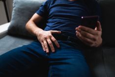 Penis Masturbation Abbildung eines männlichen Körpers abwärts des Kopfes, welcher in blauer Jeans und blauem T-Shirt auf einem grauen Sofa sitzt und auf ein Smartphone in seiner linken Hand schaut, welche in Selfie-Manier über seinem Körper hält.