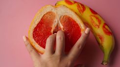 Frau führt 2 Finger in eine halbe Orange ein, daneben liegt eine Banane mit Lippenstiftabdrücken