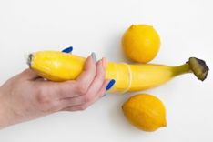 Hand hält eine Banane, über die ein Kondom gezogen ist, zwei Zitronen symbolisieren die Hoden