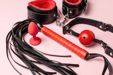 rot-schwarzes BDSM-Set mit Peitsche Knäbel und Handschellen auf rosa Untergrund