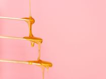 Drei Holzstäbchen an denen Honig herunterläuft vor rosanem Hintergrund