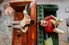 Zwei Sofas stehen mit der Rückenlehne aneinander. Auf dem linken Sofa sitzt eine Frau, auf dem rechten Sofa ein Mann. 