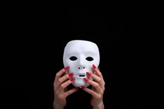 Hände mit roten langen Nägeln halten eine weiße Maske vor schwarzem Hintergrund