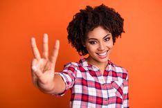 Frau mit braunen Haaren und kariertem Hemd, die drei Finger mit ihrer Hand in die Kamera zeigt vor orangem Hintergrund