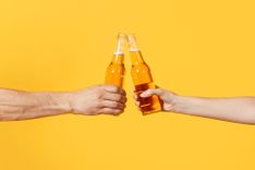 Zwei Hände vor einem gelben Hintergrund die mit Bier anstoßen