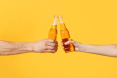 Zwei Hände vor einem gelben Hintergrund die mit Bier anstoßen
