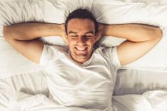 Mann mit weißem Shirt, der seine Arme hinter den Kopf verschränkt, lacht und im Bett unter der Decke liegt