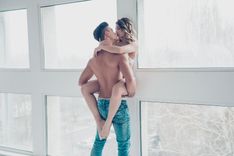 Oberkörperfreier Mann in Jeans, der eine Frau in weißer Unterwäsche trägt, gegen eine Wand drückt und küsst