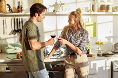Mann in grünem Shirt und blauer Jeans und eine Frau in kariertem Hemd und brauner Hose, die in einer Küche stehen und mit einem Rotwein anstoßen