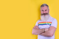 Eine Person vor einem gelben Hintergrund, mit lächelndem Gesicht, trägt ein T-Shirt mit einem LGBTIQ-Regenbogenherz-Design.