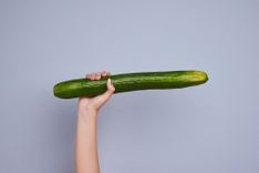 Frauenhand, die eine grüne Gurke in die Luft hält vor grauem Hintergrund