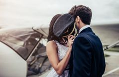 Ein Pärchen das vor einem Flugzeug steht und sich dabei mit einer vorgehaltenen Piloten-Mütze küsst