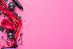 Schwarzer Knebel, schwarzer Dildo, Nippelklemmen, rote BDSM-Fesseln und eine rote Peitsche links auf rosanem Untergrund