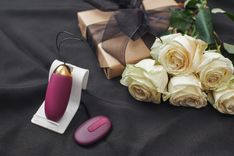 Bordeaux Vibro-Ei mit Fernbedienung auf einem schwarzen Tuch mit weißen Rosen und einem goldenen Geschenk mit schwarzer Schleife daneben