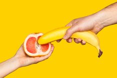 Hand, die eine halbe Grapefruit hält und eine weitere Hand, die eine Banane in die Grapefruit steckt vor gelbem Hintergrund