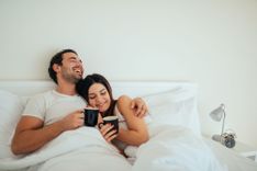 Paar liegt mit Kaffeetassen im Bett und lacht