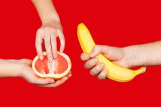 Eine Hand, die eine Banane hält und zwei weitere Hände, die eine halbierte Grapefruit halten vor rotem Hintergrund