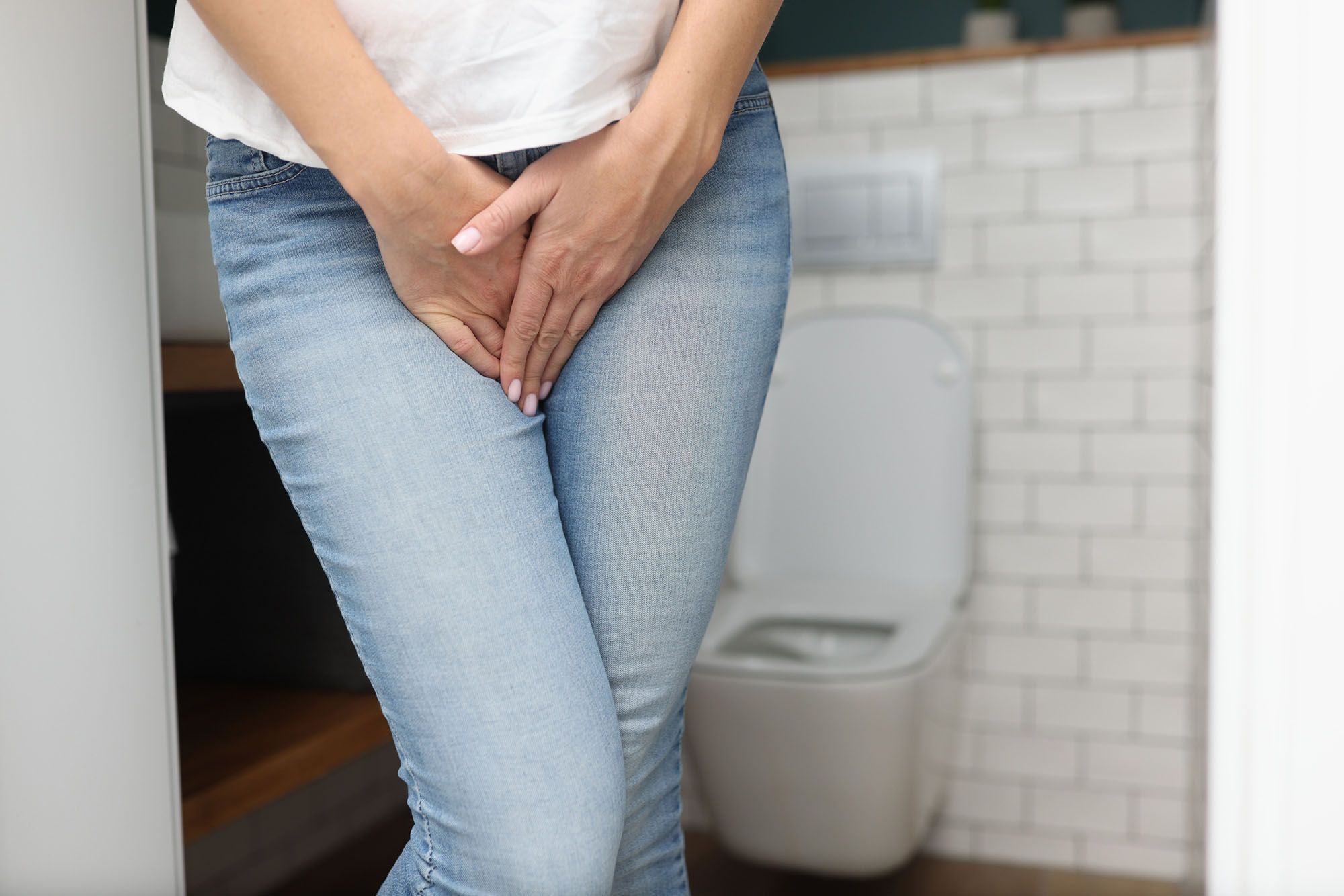 Frau in Jeans und weißem T-Shirt, die sich beide Hände vor den Schritt hält vor einer Toilette im Hintergrund