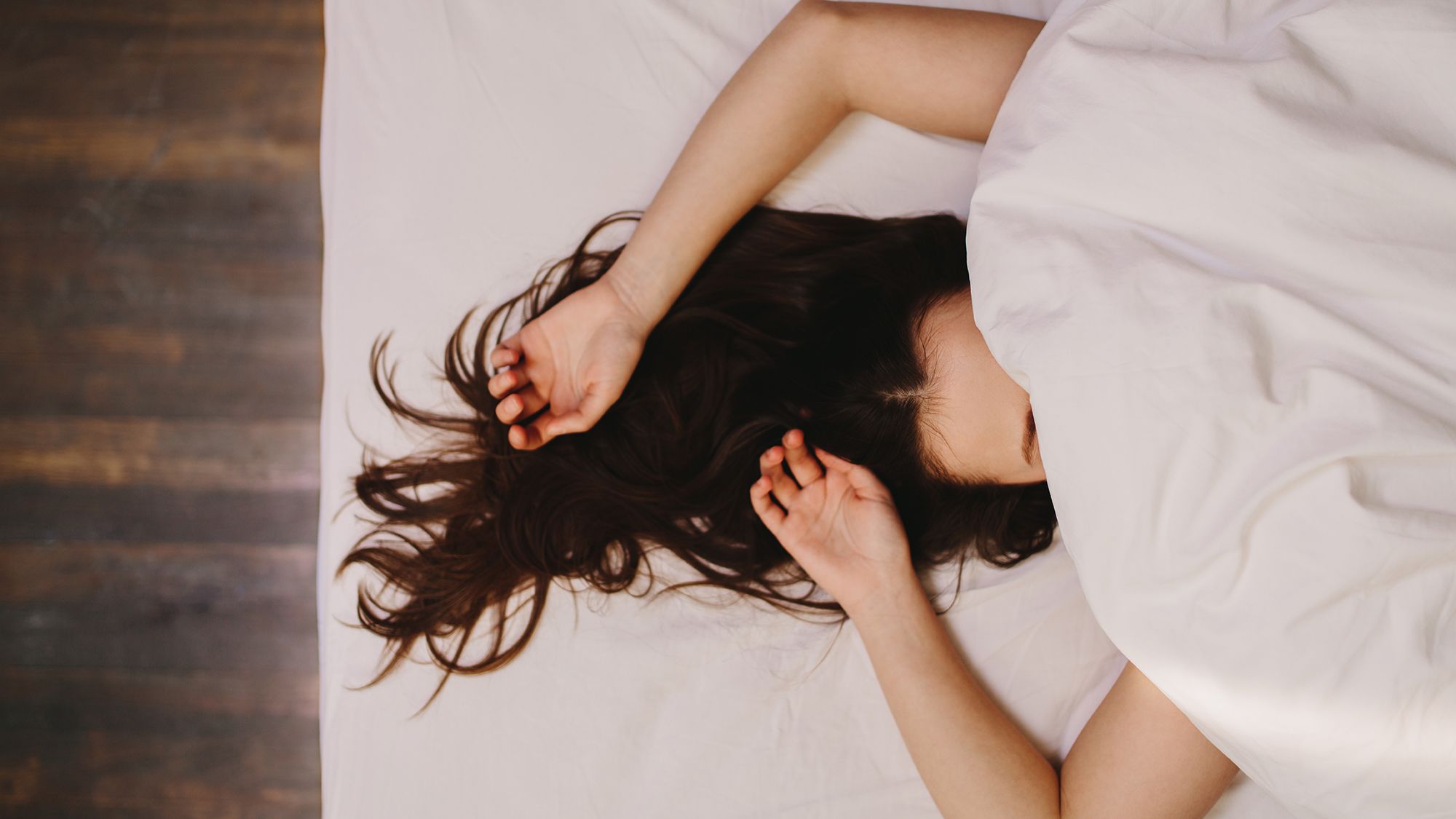 Brünette Frau unter einer weißen Decke auf einem Bett, die ihren halben Kopf und ihre Arme nach oben streckt