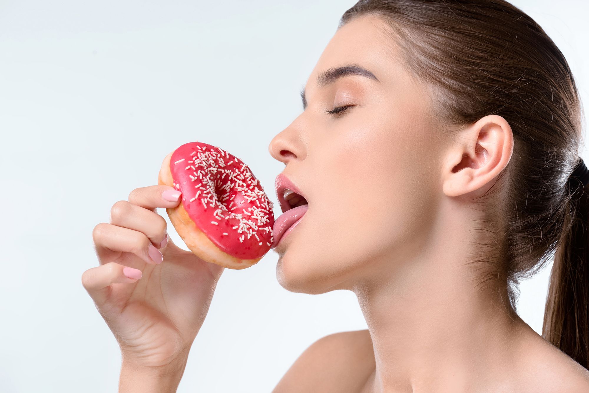Frau leckt lustvoll an einem pinkglasierten Donut mit Streuseln