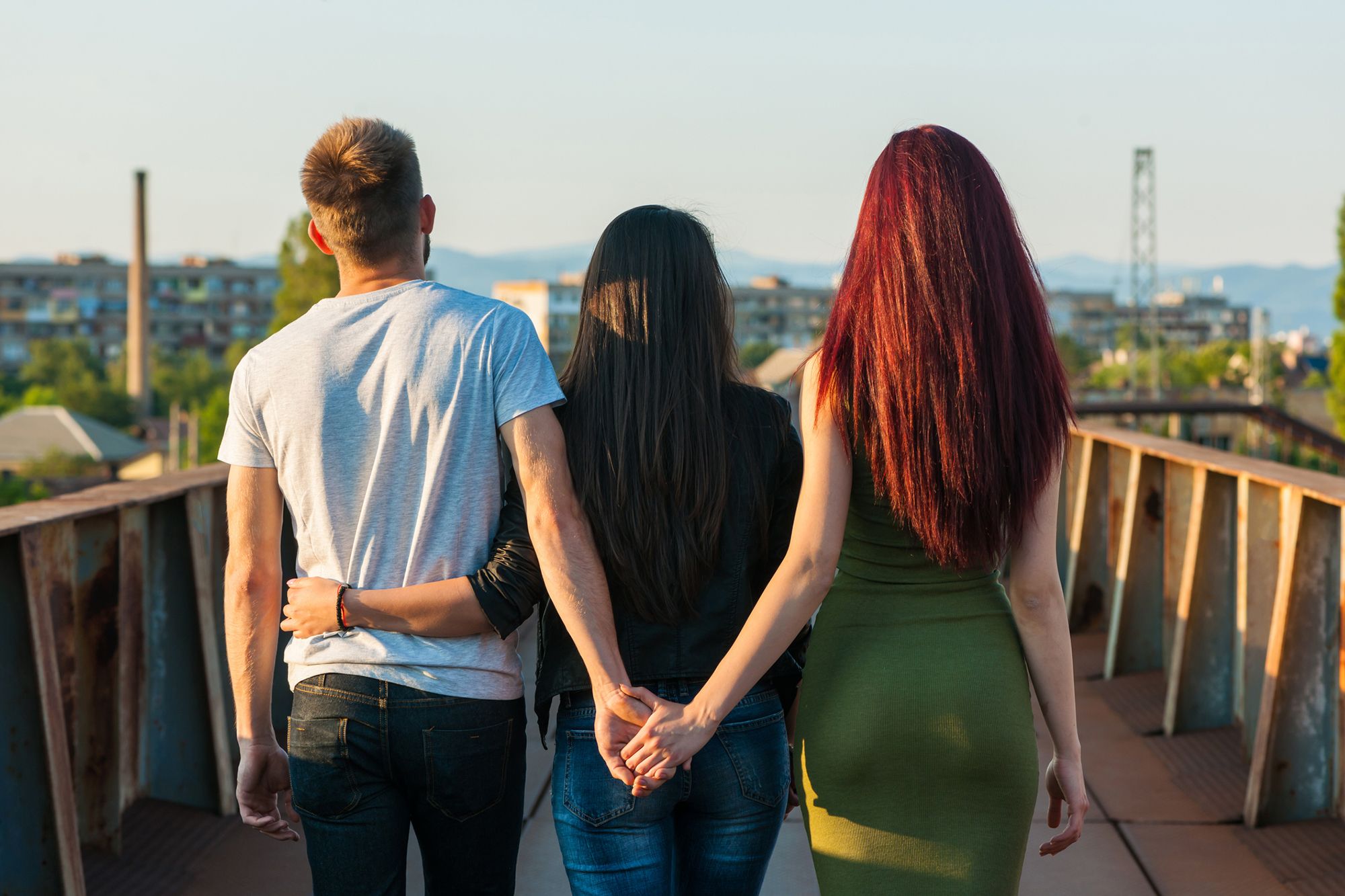 Eine rothaarige Frau in einem grünen Kleid und ein Mann in weißem Shirt und schwarzer Jeans, die Händchen hinter dem Rücken einer schwarzhaarigen Frau in schwarzem Blazer und blauer Jeans halten, die eine Hand um die Hüfte des Mannes schlingt